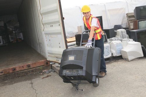 A female worker wearing a yellow safety helmet, high-visibility vest, and gloves is standing outside a storage container, lifting and carrying a black computer monitor. The monitor appears large, with visible cables hanging from it. Behind her, there are various electronic items, including old desktop computers and computer towers, stacked or placed against a white wall or fence, suggesting a collection or clearance of electronic waste. The ground is paved, with some minor cracks visible, and the environment appears to be an outdoor waste disposal or recycling yard, possibly in the Waltham Forest area, where Garden Clearance Waltham Forest offers rubbish removal services. The scene captures the process of clearing or disposing of electronic waste materials, with the worker actively handling the items as part of a clearance operation.
