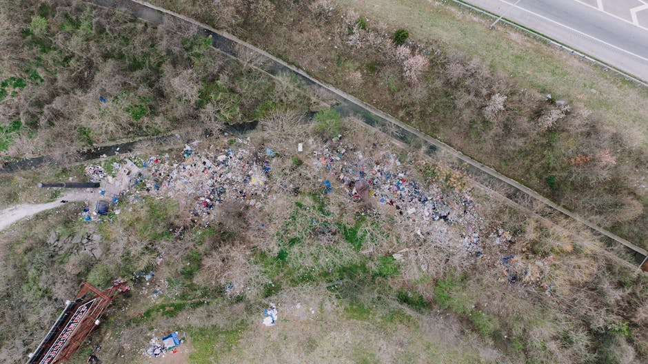 A small pile of mixed waste, including plastic bottles, paper, and other debris, is settled on a muddy, uneven shoreline next to a body of water. The waste is scattered over a patch of damp soil with 