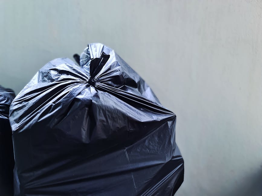 Two large black plastic garbage bags, filled and tied at the top, are positioned on the edge of a sidewalk in an outdoor urban setting. The bags are placed next to a low curb, with one slightly leanin
